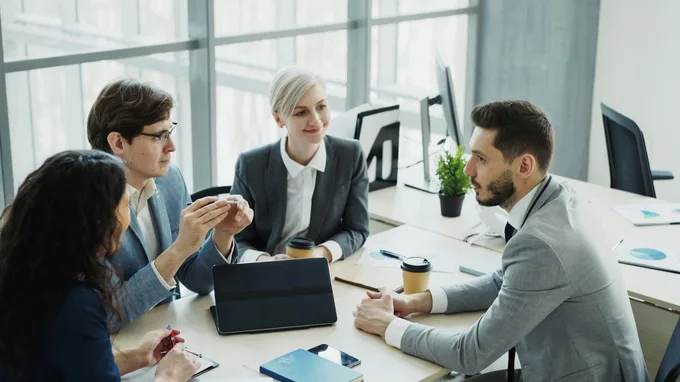 Four colleagues around a table working something through together — one explaining, another taking notes